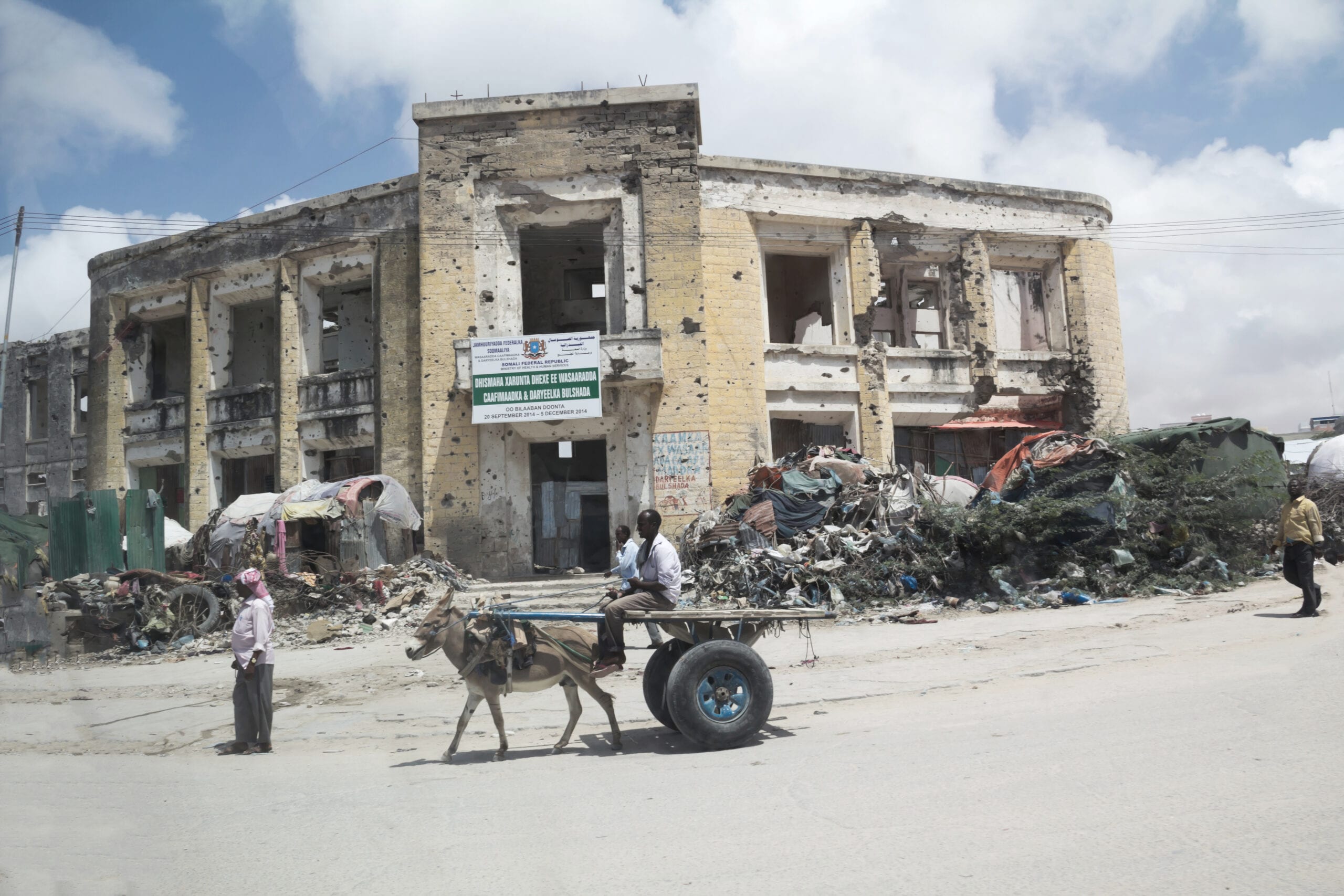 A bullet-scarred yellow ministry building in Mogadishu, Somalia, with makeshift shelters beside it and a donkey cart passing in front, 2014.